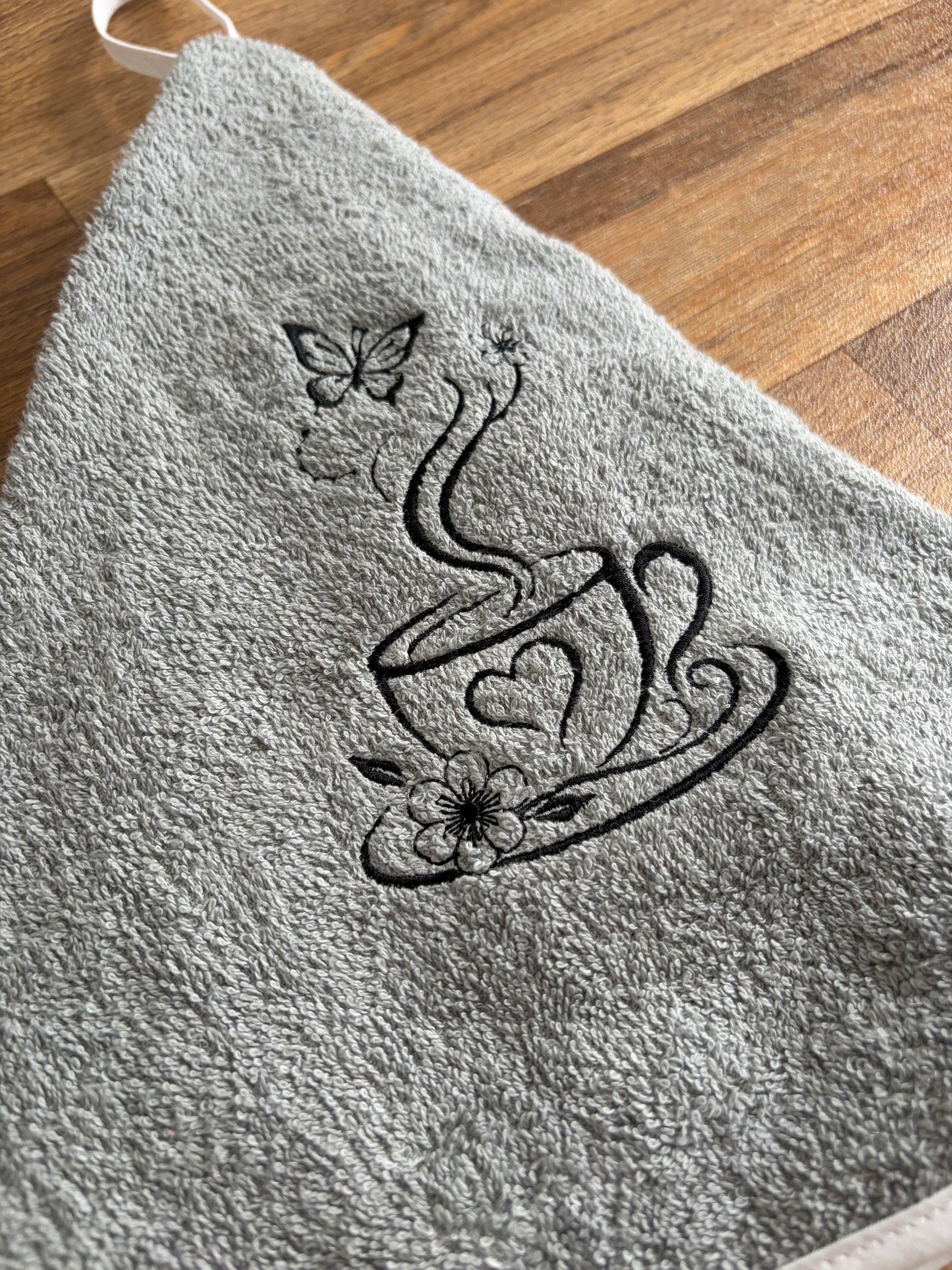 Détail de la broderie noire sur éponge grise : tasse ornée d’un cœur, accompagnée de fleurs et d’un papillon