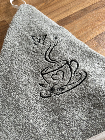 Détail de la broderie noire sur éponge grise : tasse ornée d’un cœur, accompagnée de fleurs et d’un papillon