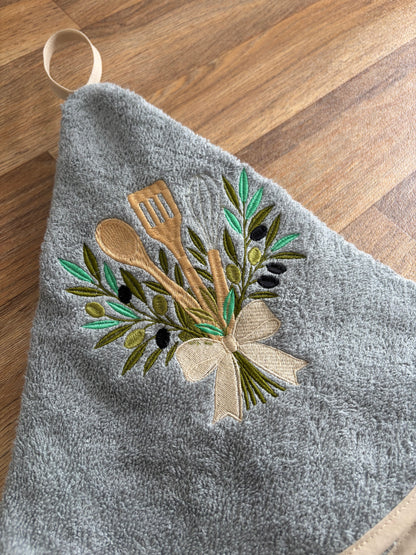Gros plan sur la broderie d’ustensiles de cuisine entourés de branches d’olivier et réunis par un nœud écru