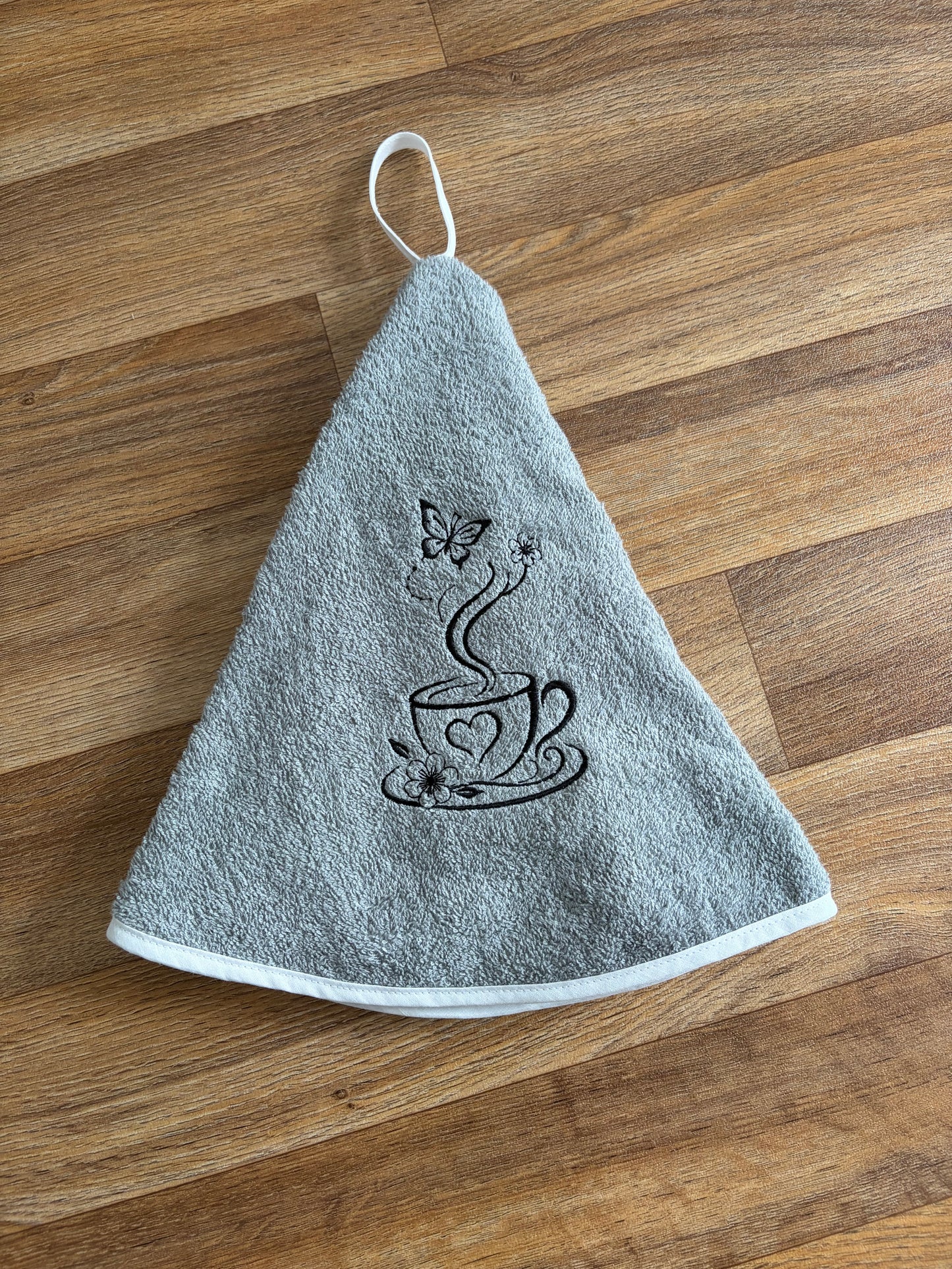 Essuie-mains rond gris avec broderie noire représentant une tasse, des fleurs et un papillon, et bordure blanche
