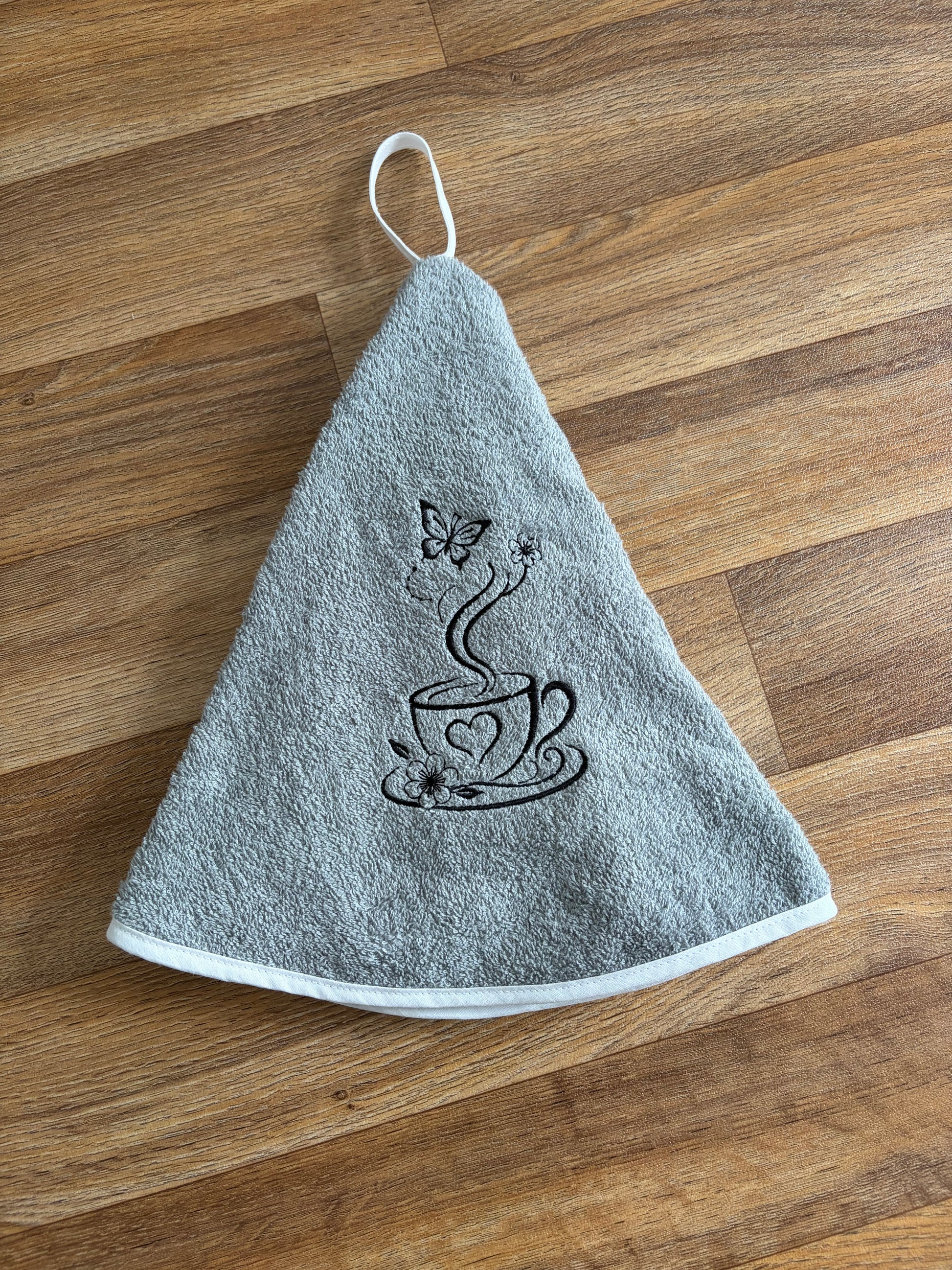 Essuie-mains rond gris avec broderie noire représentant une tasse, des fleurs et un papillon, et bordure blanche