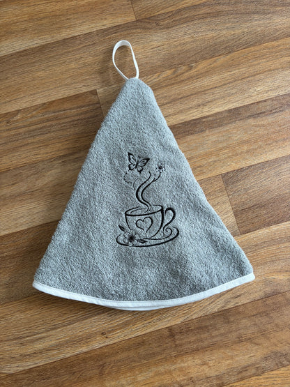 Essuie-mains rond gris avec broderie noire représentant une tasse, des fleurs et un papillon, et bordure blanche